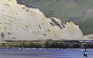 big whitish cliff face at edge of beach with person in blue shirt and black pants running into the water